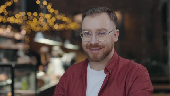 Portrait View of the Cheerful Bearded Male Student Wearing Glasses Looking at Camera Sitting in Cafe alt
