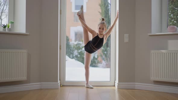 Wide Shot Flexible Talented Teenage Gymnast Stretching One Leg Up Looking at Camera Standing Indoors alt