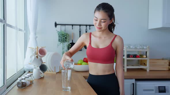 Portrait of Asian attractive sport woman pour water into glass after workout exercise in kitchen. alt