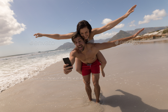 Happy young couple piggyback with arms outstretched taking selfie with mobile phone at beach - Stock Photo - Images