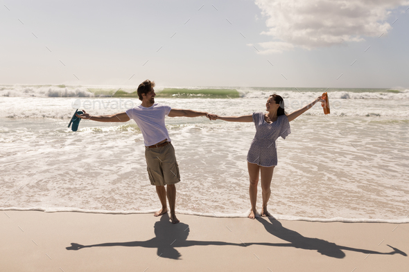 Romantic Happy Young Couple With Arms Outstretched And Holding Hands On Beach In The Sunshine Stock Photo By Wavebreakmedia