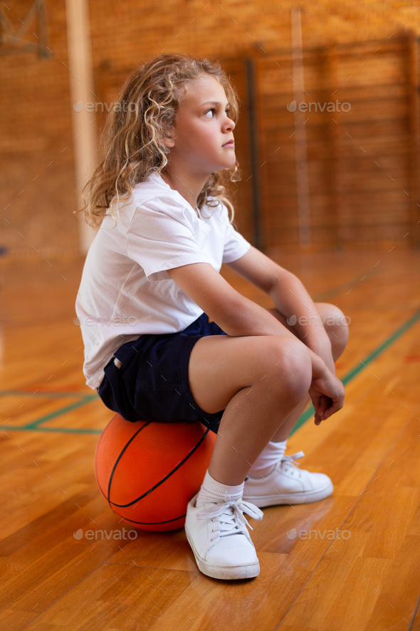 Side view of a mixed -race schoolgirl sitting on a basketball in basketball court at school