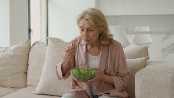 Beautiful Elderly Woman Eats Her Vegetable Salad Sitting at Home on the Couch alt