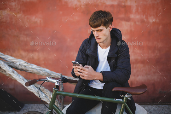 Young man in down jacket and white t-shirt sitting with mobile phone in hands and bicycle nearby - Stock Photo - Images
