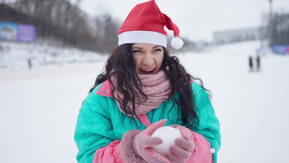 Portrait of Joyful Woman in Christmas Hat Playing Snowballs at Ski Resort alt