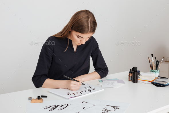 Portrait of young beautiful lady sitting at the white desk and writing alphabet on paper isolated
