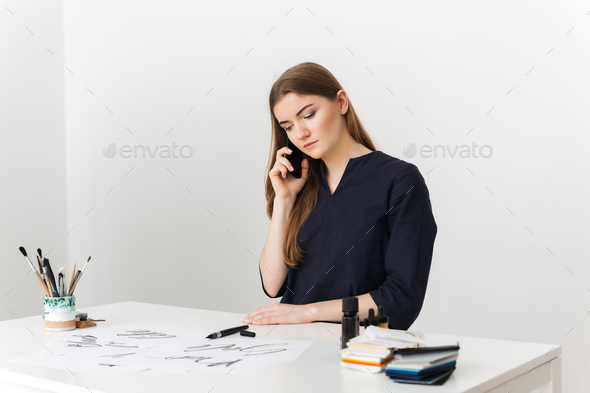 Portrait of young pretty lady sitting at the white desk and talking on her cellphone - Stock Photo - Images