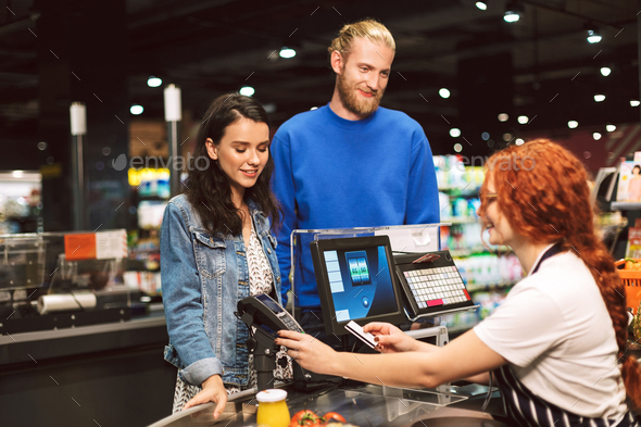 Young smiling couple standing near cashier desk while happily pa