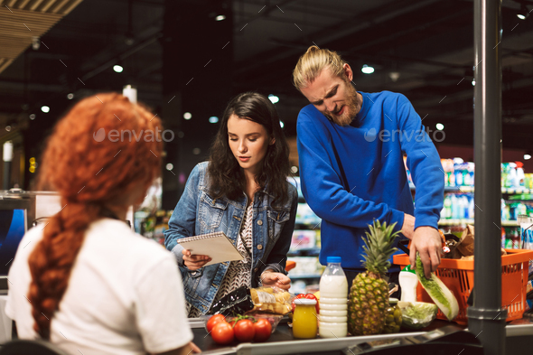 Young woman with shopping list standing near cashier desk while