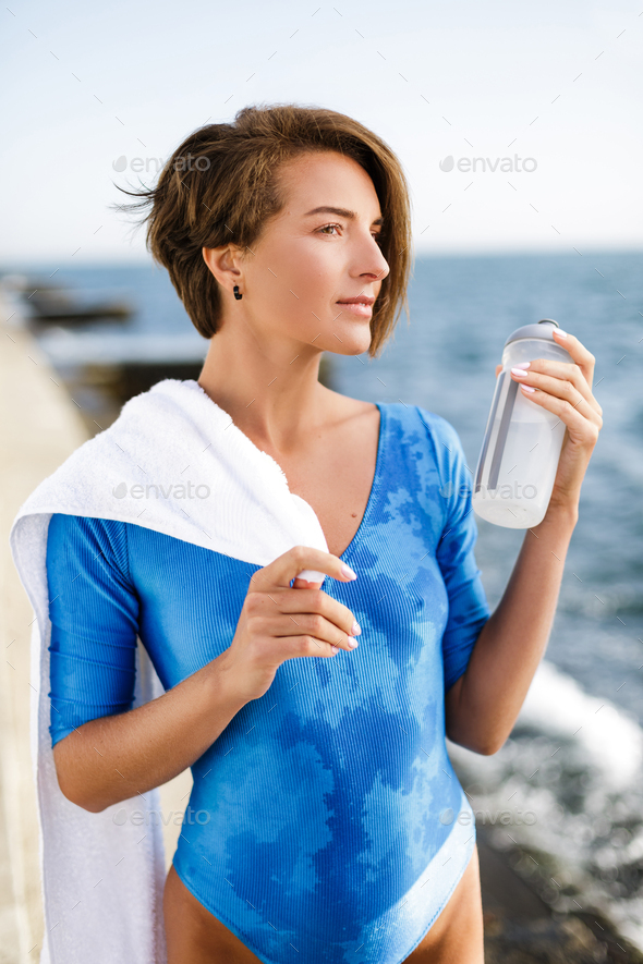 Portrait of pretty woman in blue swimsuit standing with white towel on shoulder and bottle in hand - Stock Photo - Images