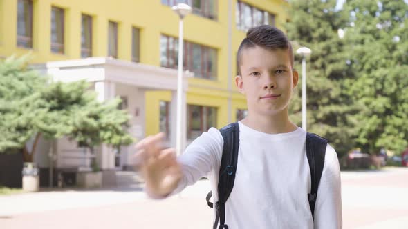 A Caucasian Teenage Boy Waves at the Camera with a Smile  a School in the Background