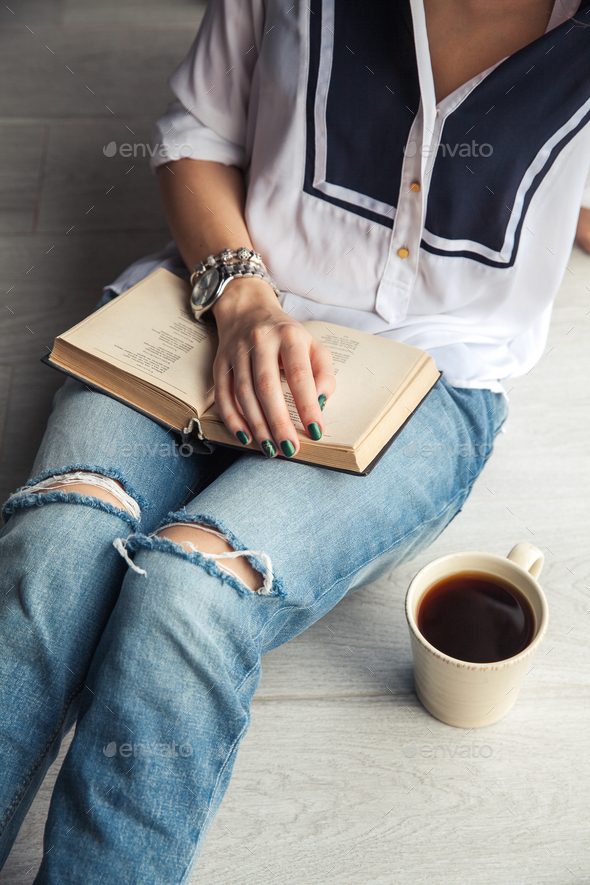 Young modern girl in torn jeans reading a book with a big cup of coffee. Fashion, lifestyle