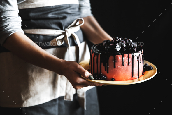 Woman Chef in black apron holds cake decorated with berries and fruits with two hands