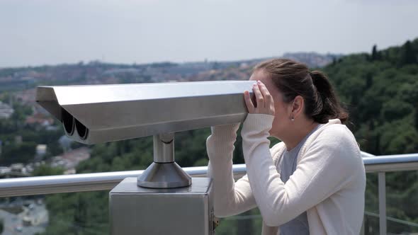 Woman Looks Through Binoculars Exploring Cityscape Along Bay alt