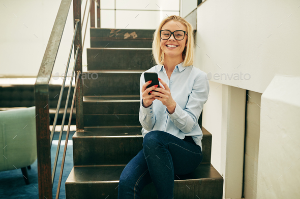 Laughing businesswoman sitting on office stairs reading a funny text - Stock Photo - Images