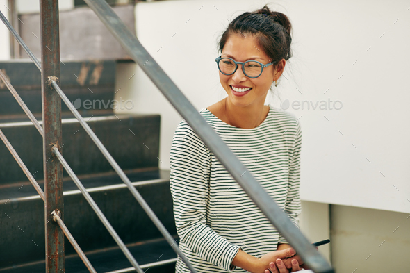 Laughing Asian businesswoman sitting on office stairs reading text messages
