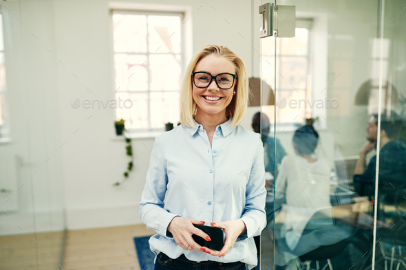 Smiling young businesswoman holding her cellphone in an office - Stock Photo - Images