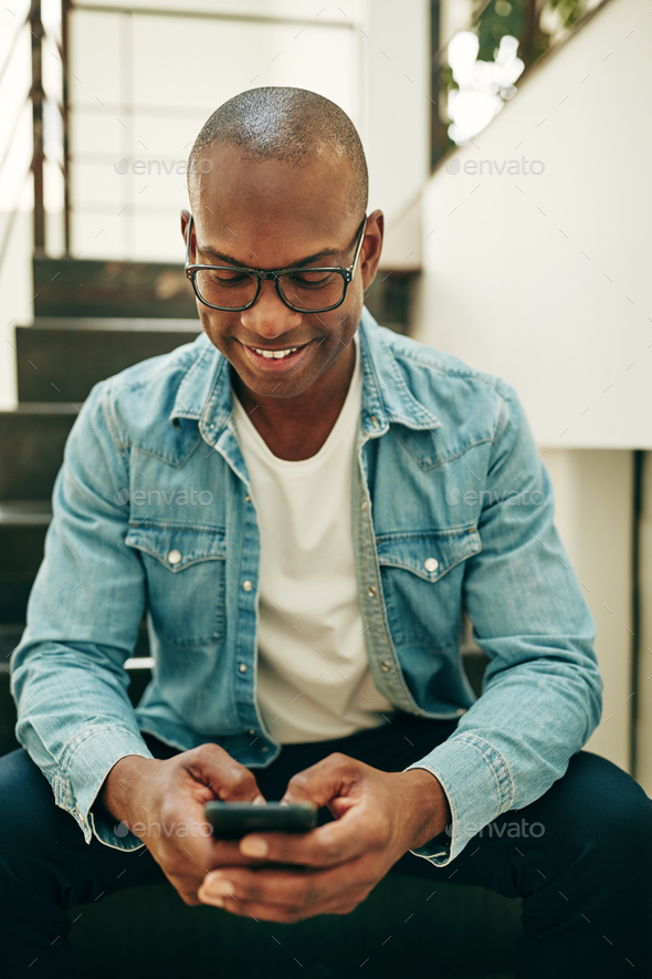 Smiling young African businessman sitting on office stairs text messaging - Stock Photo - Images