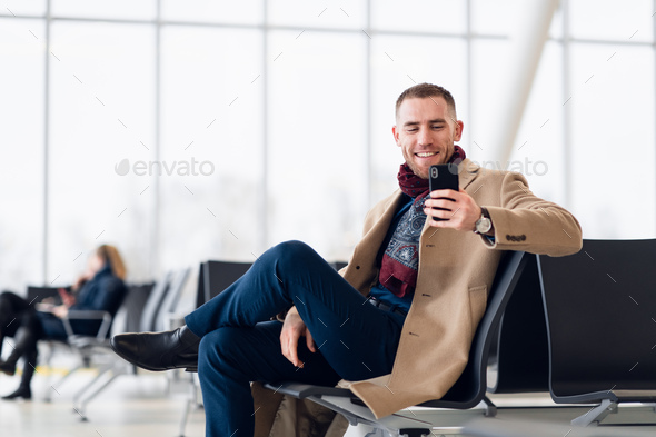 Attractive young adult man in stylish scarf and coat sitting inside airport terminal waiting area - Stock Photo - Images