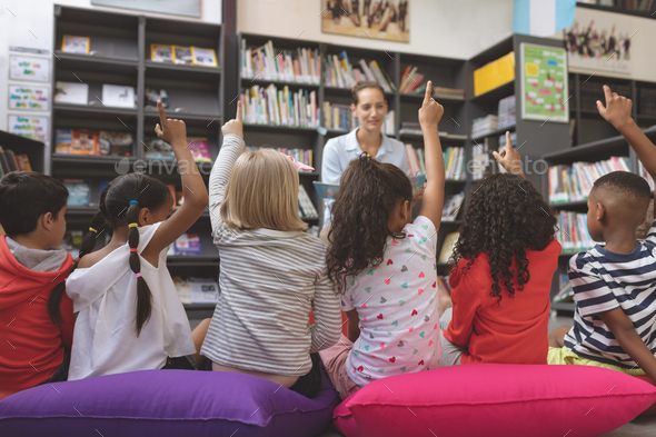 Schoolkids raising hand to answer at a question asking agaisnt bookshelf - Stock Photo - Images