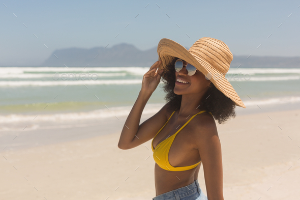 Happy young African American woman in yellow bikini, hat and sunglasses  standing on the beach Stock Photo by Wavebreakmedia