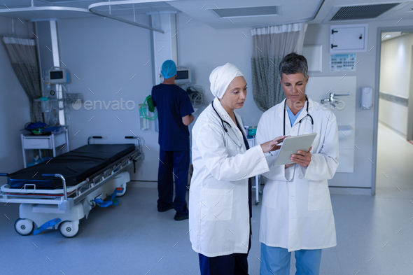 Female doctors discussing over digital tablet in clinic while a doctor is working behind them - Stock Photo - Images