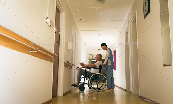 Nurse pushing disabled senior male patient sitting in wheelchair in corridor at retirement home
