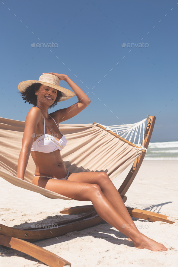 Woman in white bikini with hat sitting on hammock while smiling at the camera at beach