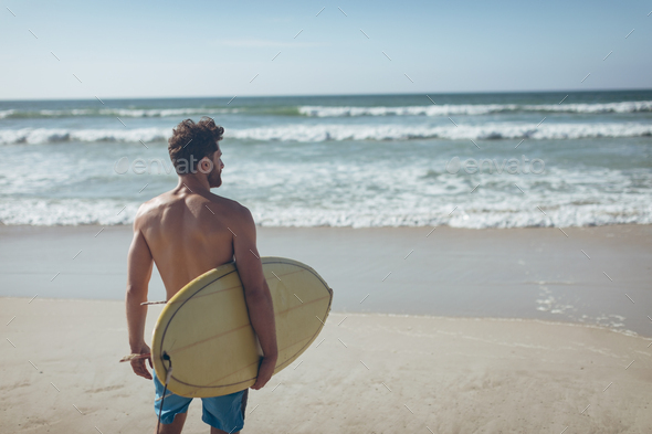 Young male surfer with a surfboard standing on a beach on sunny day. He is looking the waves