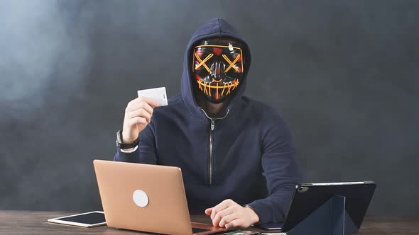 Portrait of a Masked Male Hacker with a Laptop and Credit Card in Hand. alt