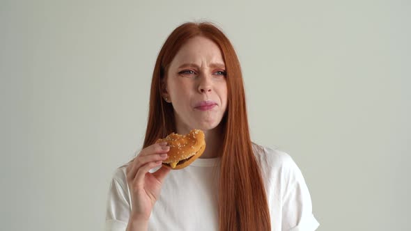 Closeup Portrait of Hungry Young Woman Eating Unappetizing Hamburger Dissatisfied with Food Quality alt