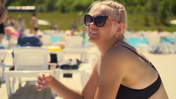 Slow Motion Closeup Blonde Girl Sits on Deckchair on Beach in Sunglasses Touches Her Hair Which Is alt