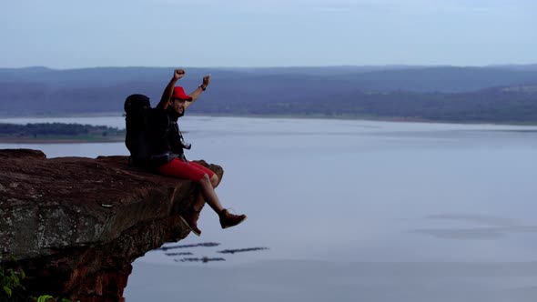 cheerful hiker man sitting and gesture raised arms on the edge of cliff alt