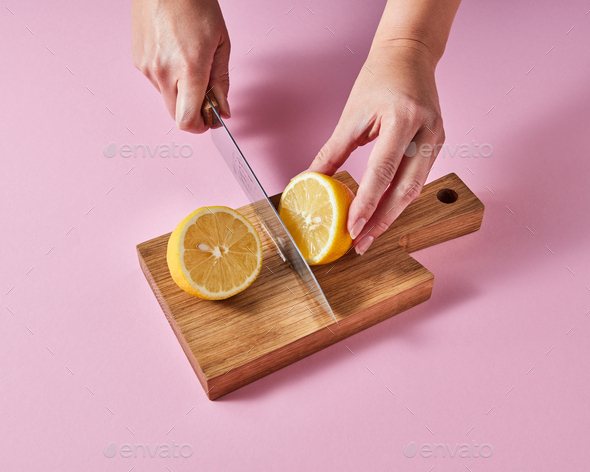 Woman&#39;s hands cut a lemon on a wooden board with a knife around a pink background with copy - Stock Photo - Images