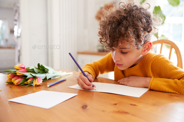 Young Boy At Home With Bunch Of Flowers Writing In Mothers Day Card
