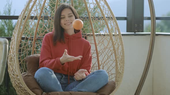 Smiling Young brunette woman in red hoodie with orange sitting in hanging chair on home terrace alt