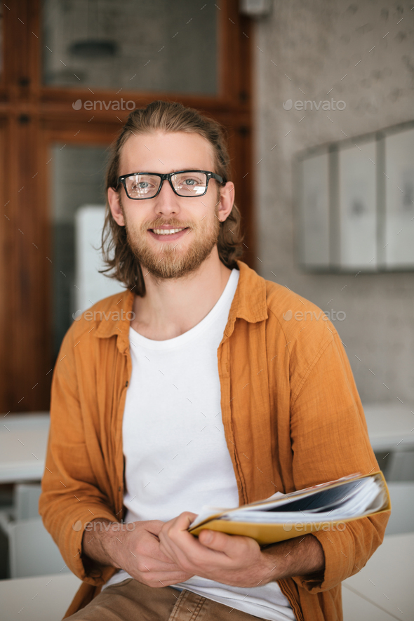 Portrait of young smiling man sitting on desk in auditorium with document case in hands - Stock Photo - Images