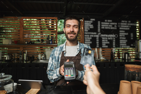 Portrait of affable barista man taking credit card from customer