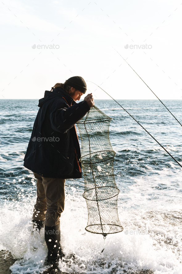 Handsome young man fisherman wearing coat and hat at the seashore.