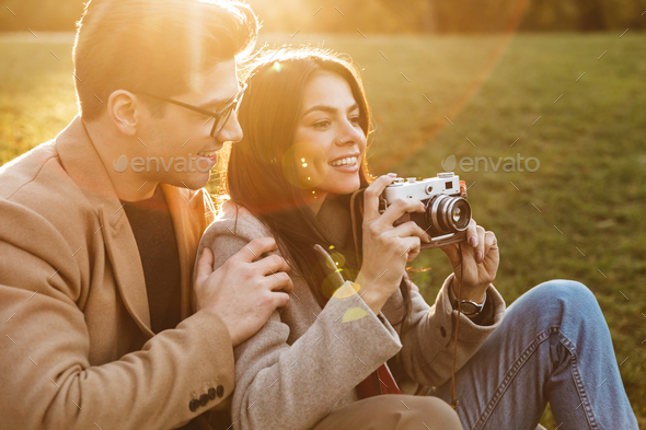 Portrait of casual couple aking photo on retro camera and smiling while sitting on grass in park
