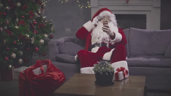 Man in Santa Claus Costume Taking Selfie on Smartphone Sitting on Couch alt