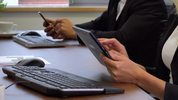 Two office workers, sits at desks in front of computers and work on a tablet and a smartphone alt