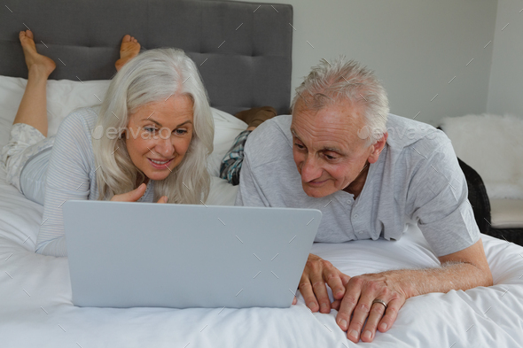 Front view of active senior Caucasian couple using laptop while lying on bed in bedroom at home