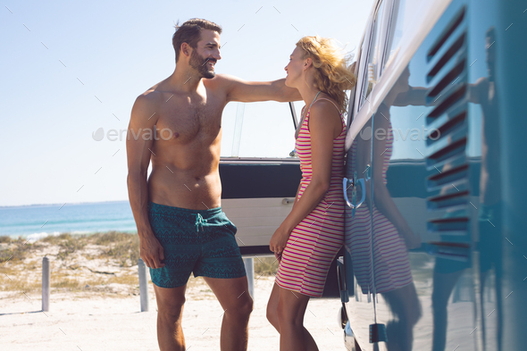 Side view of young Caucasian couple talking with each other near camper van at beach in the sunshine