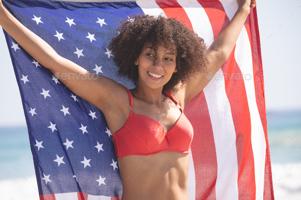 Front view of happy African american woman in bikini holding american flag on the beach
