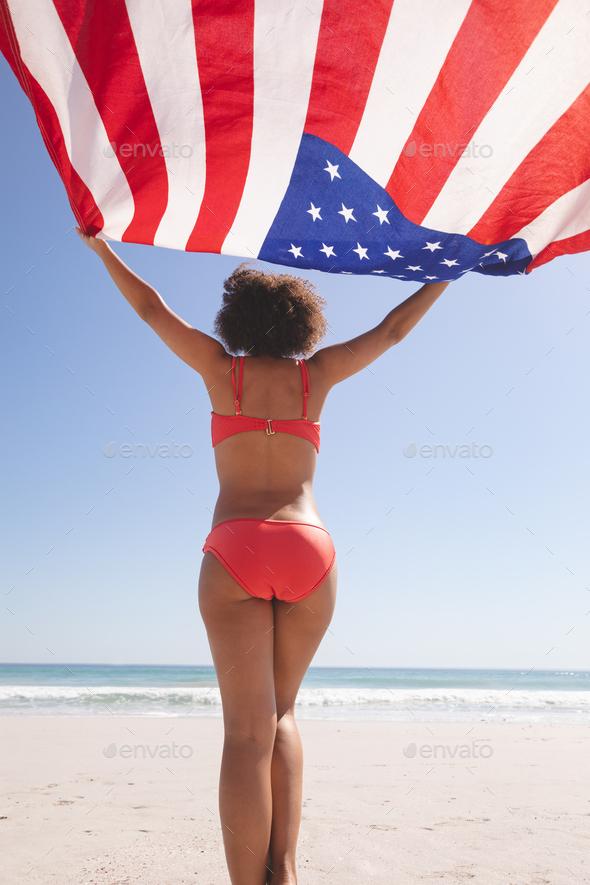 Rear view of African american woman in bikini holding american flag on the beach