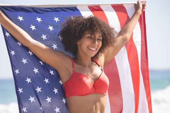 Front view of happy African american woman in bikini holding american flag on the beach