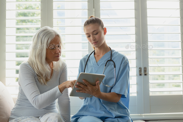 Active senior Caucasian woman and female doctor using digital tablet on window seat at home