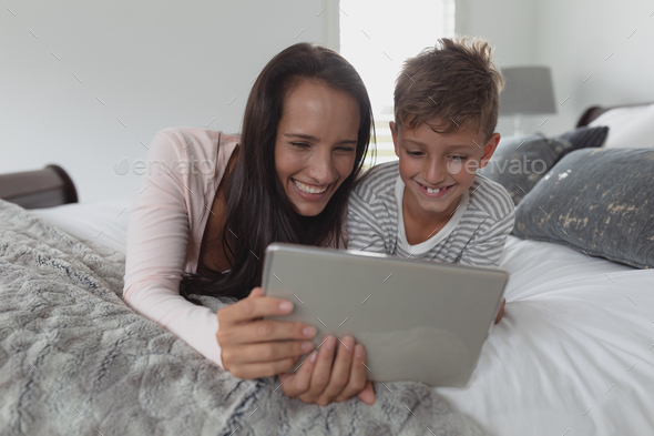 Front view of happy Caucasian mother and son using digital tablet in bedroom at home