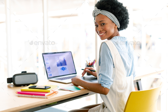 African american female graphic designer using graphic tablet while looking at laptop at desk